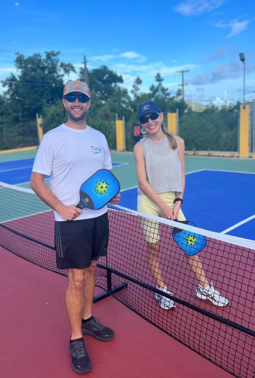 Couple leaning on the net holding matching blue paddles, smiling at the camera