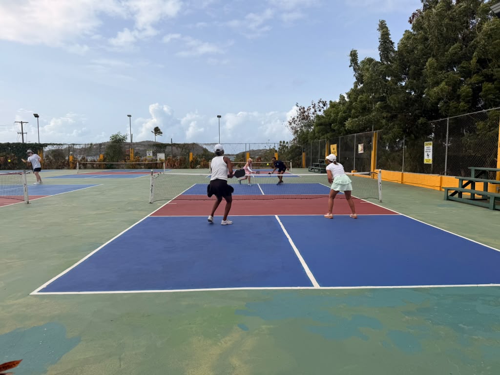 Four players mid-doubles rally across the wide court complex under blue sky