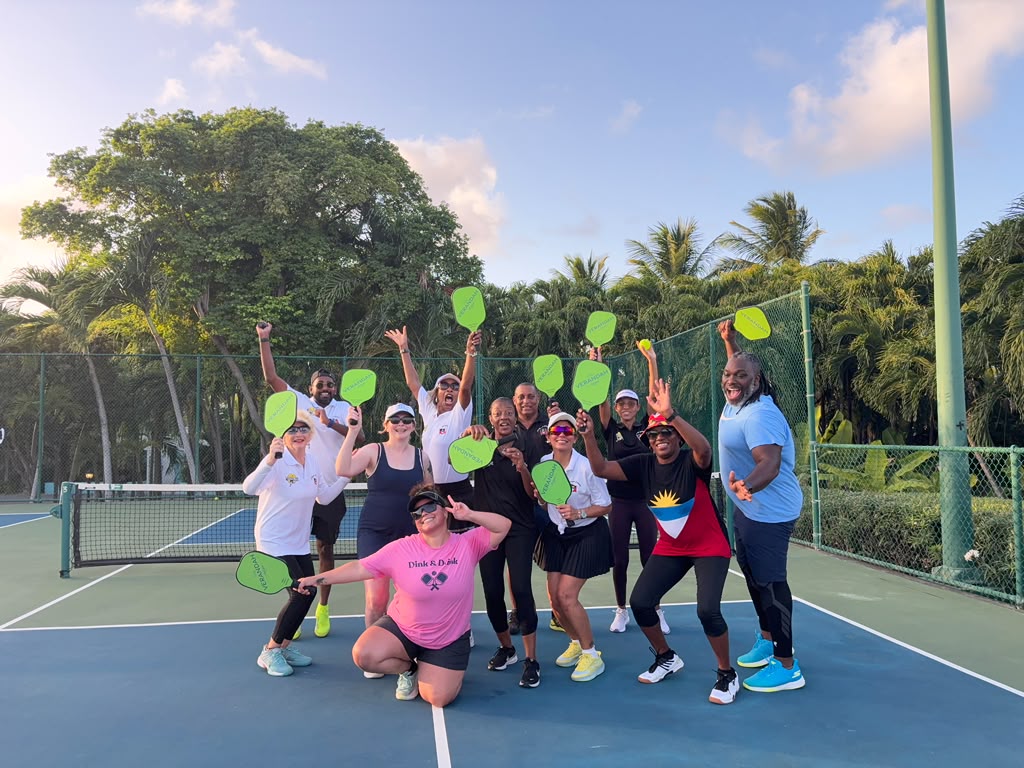 Large group holding green paddles aloft and cheering on court, palm trees behind them