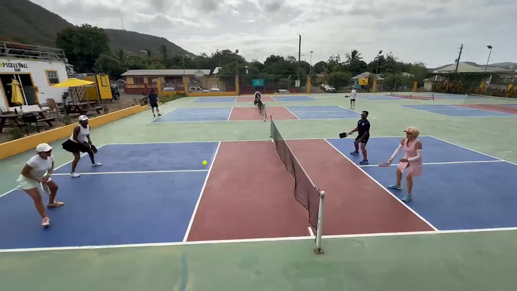Wide shot of a doubles rally in progress across the multi-court complex